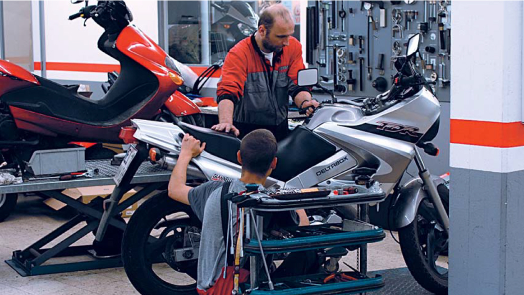 Imagine cu un atelier în care două persoane lucrează la o motocicletă.